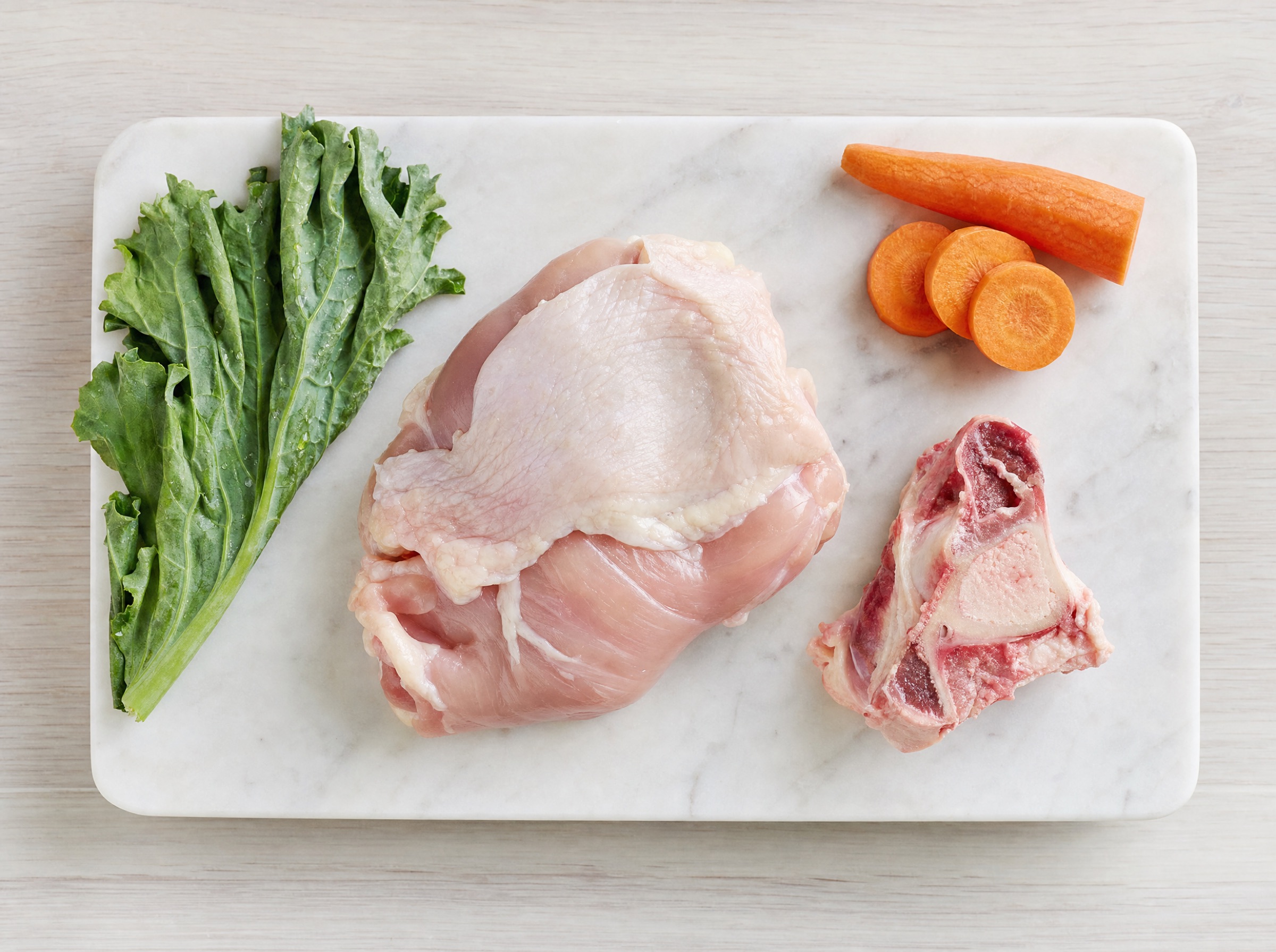 Top-down flat lay of raw dog food components: raw chicken thigh meat, raw bone, raw carrot pieces on a clean white-marble cutting board